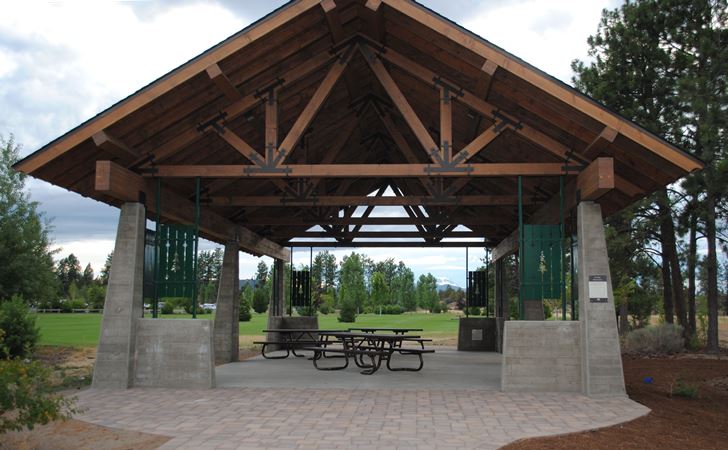 a picnic shelter at Pine Nursery Park with concrete columns, timbered beams and three empty picnic tables underneath and lawn and trees in the background