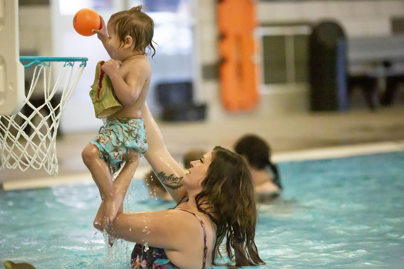 A mother lifts her child out of the pool to dunk a ball.